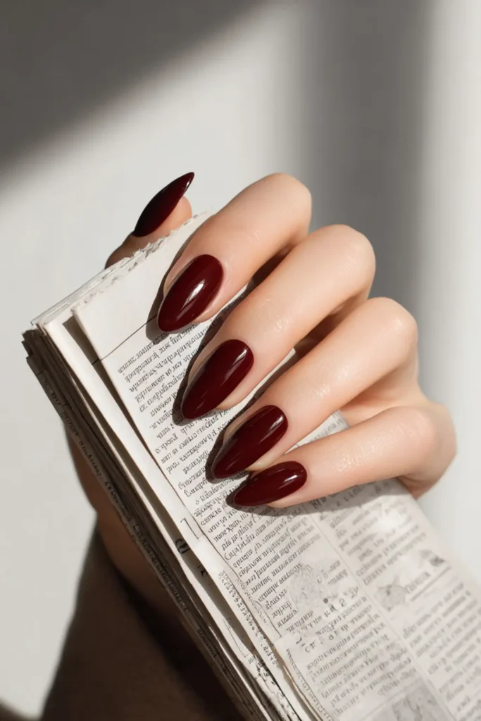 deep burgundy almond nails with a glossy finish holding a newspaper