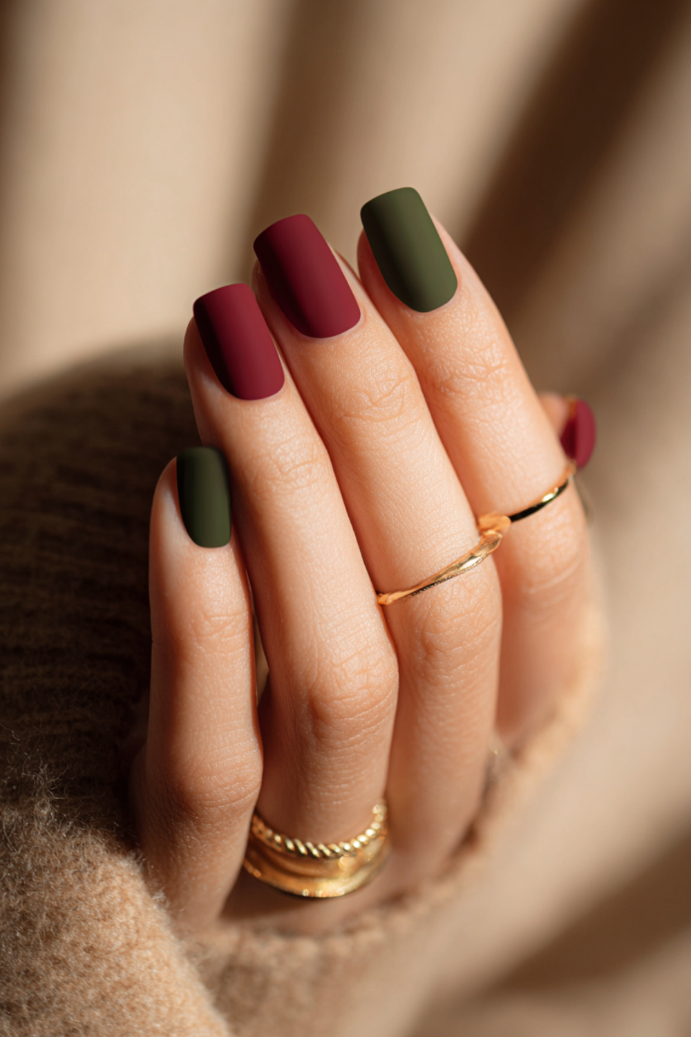 Close-up of a fall manicure with deep red and olive green matte nails, gold rings and cozy sweater background.