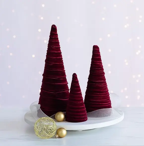 set of three burgundy velvet cone Christmas trees displayed on a white tray with gold ornaments and soft fairy lights in the background