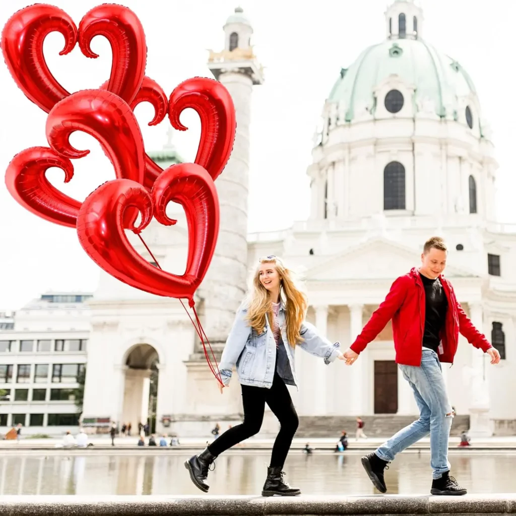 Couple holding hands with red heart balloons shaped like abstract hearts, romantic Valentine’s Day outdoor celebration.