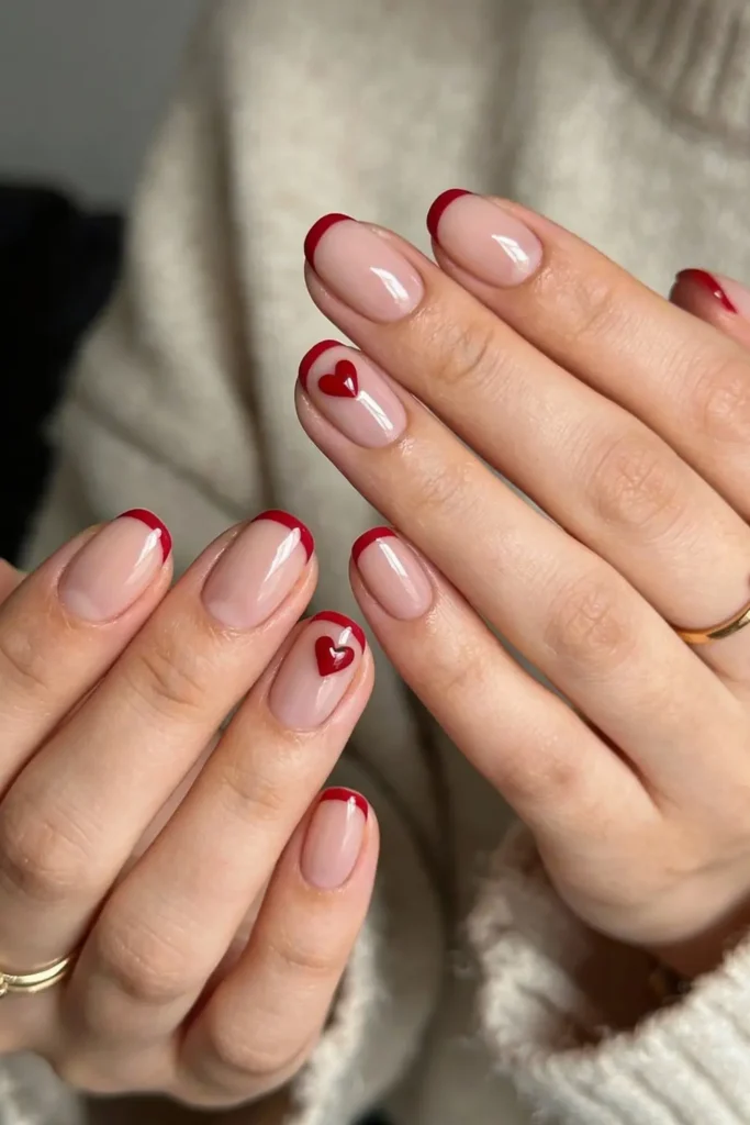 Nude French tip Valentine’s Day nails with red heart accents and glossy finish