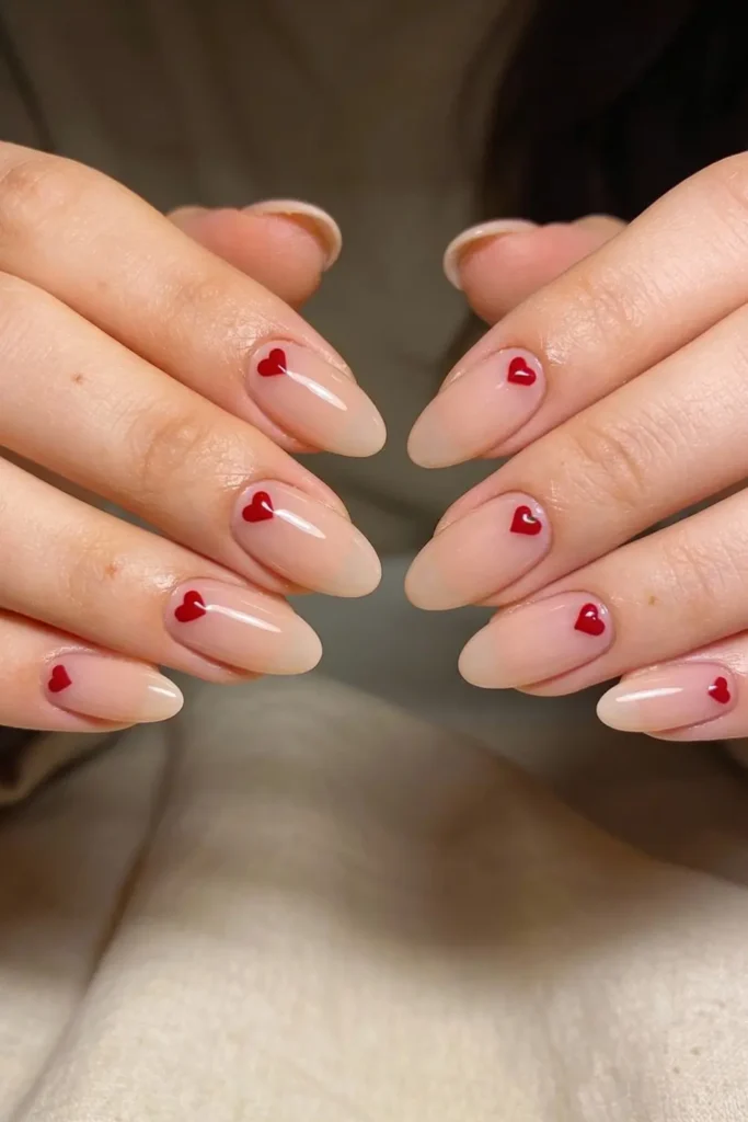 Minimal nude Valentine’s Day nails with single red heart accents and glossy finish