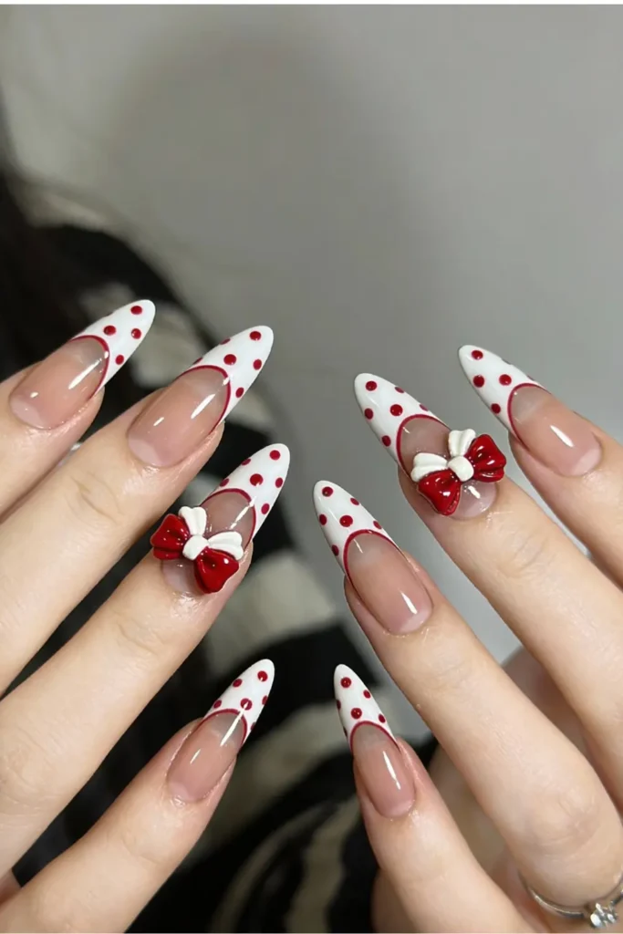White French tip Valentine’s Day nails with red polka dots and 3D bow accents on almond nails